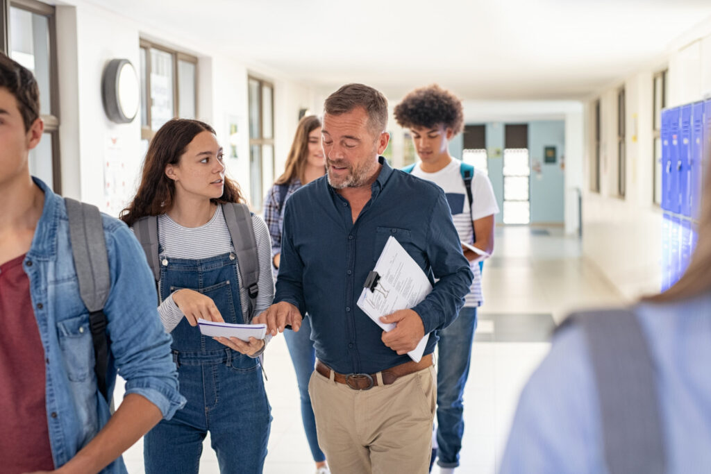 Group of students walking with a teacher in a school hallway, talking and carrying books and papers.